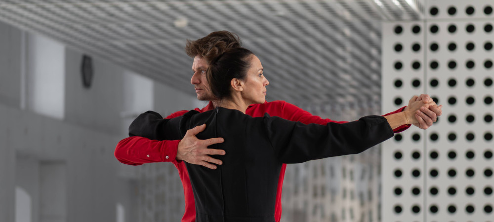 A man in a red shirt and a woman in a black shirt are standing embraced in a dance pose, each looking in their own direction.