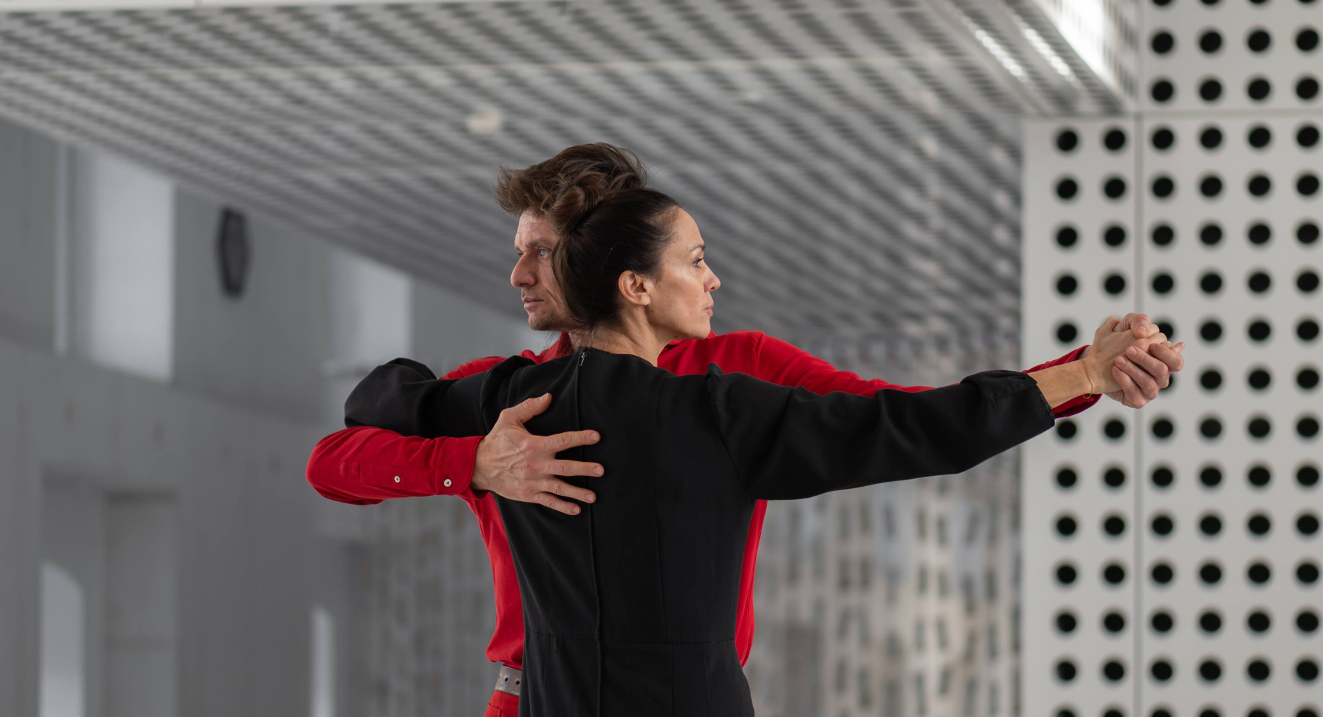 A man in a red shirt and a woman in a black shirt are standing embraced in a dance pose, each looking in their own direction.