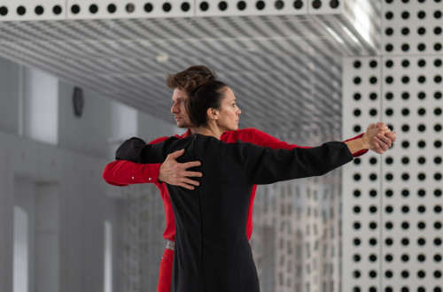 A man in a red shirt and a woman in a black shirt are standing embraced in a dance pose, each looking in their own direction.