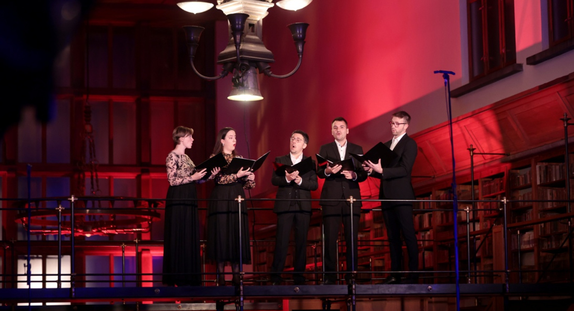 A group of singers on a high stage, with red lighting in the background.