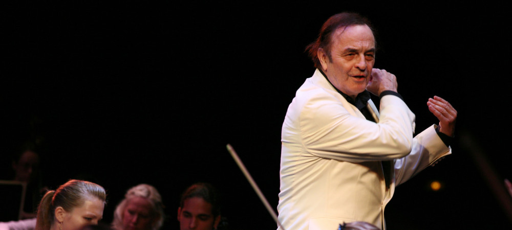 Dirigent v belem suknjicu dirigira orkester, ki je pod njim.