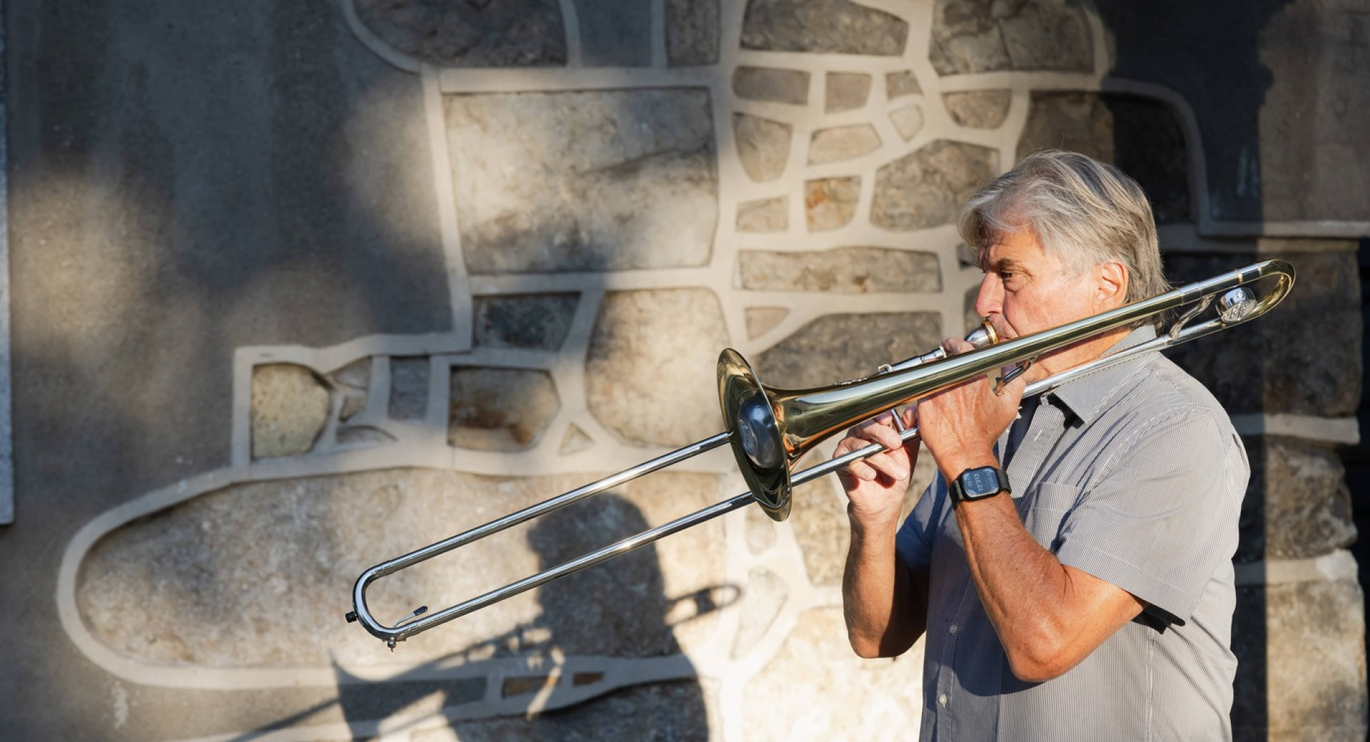 Moski v sivi srajci igra na pozavno na prostem pred kamnitim zidom, fotografiran z levega profila.