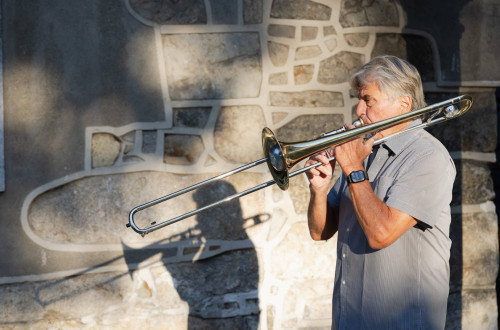 Moski v sivi srajci igra na pozavno na prostem pred kamnitim zidom, fotografiran z levega profila.