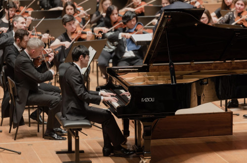 Moski v obleki sedi za klavirjem in igra nanj, za njim pa igra orkester.