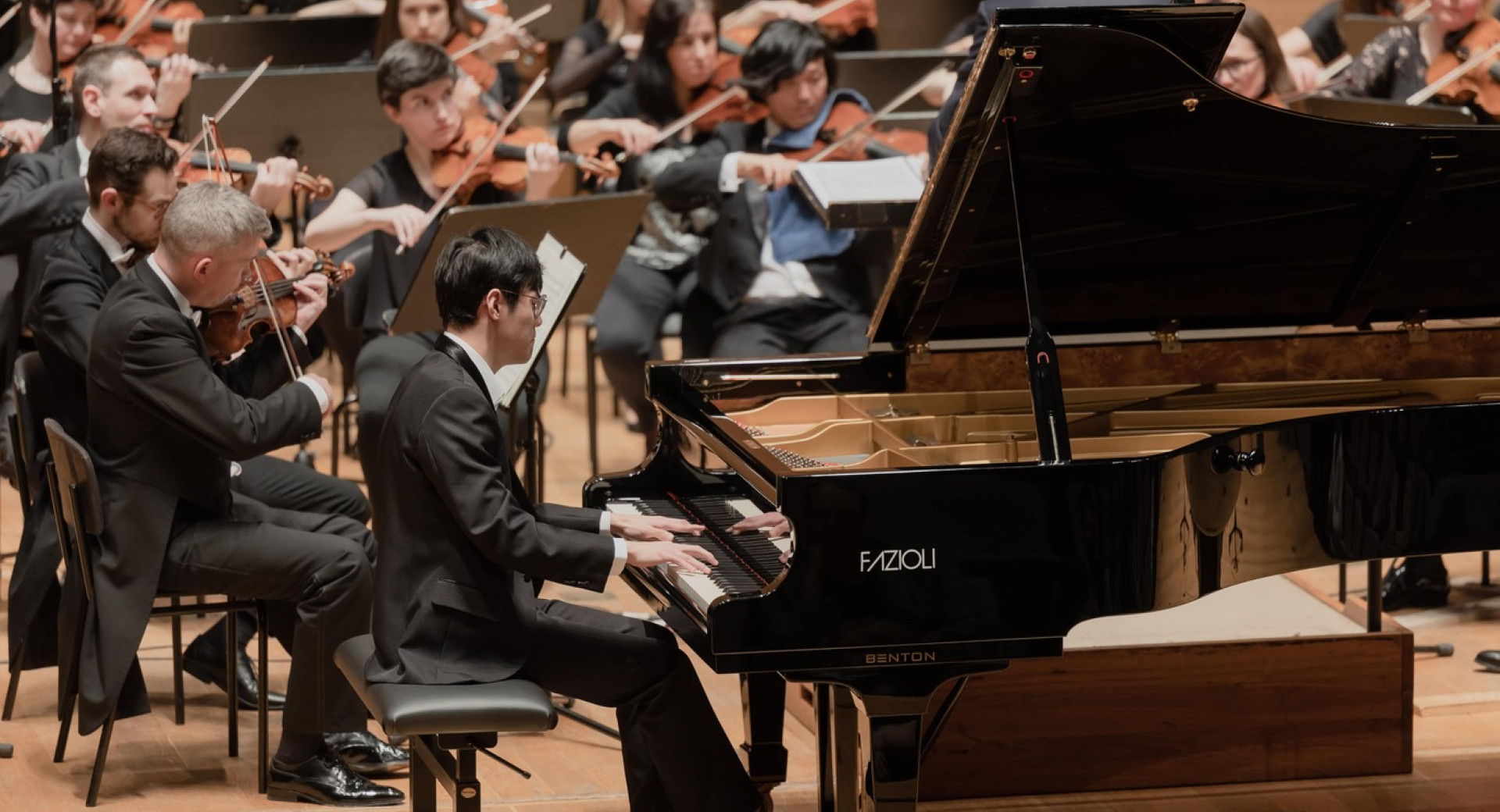 Moski v obleki sedi za klavirjem in igra nanj, za njim pa igra orkester.