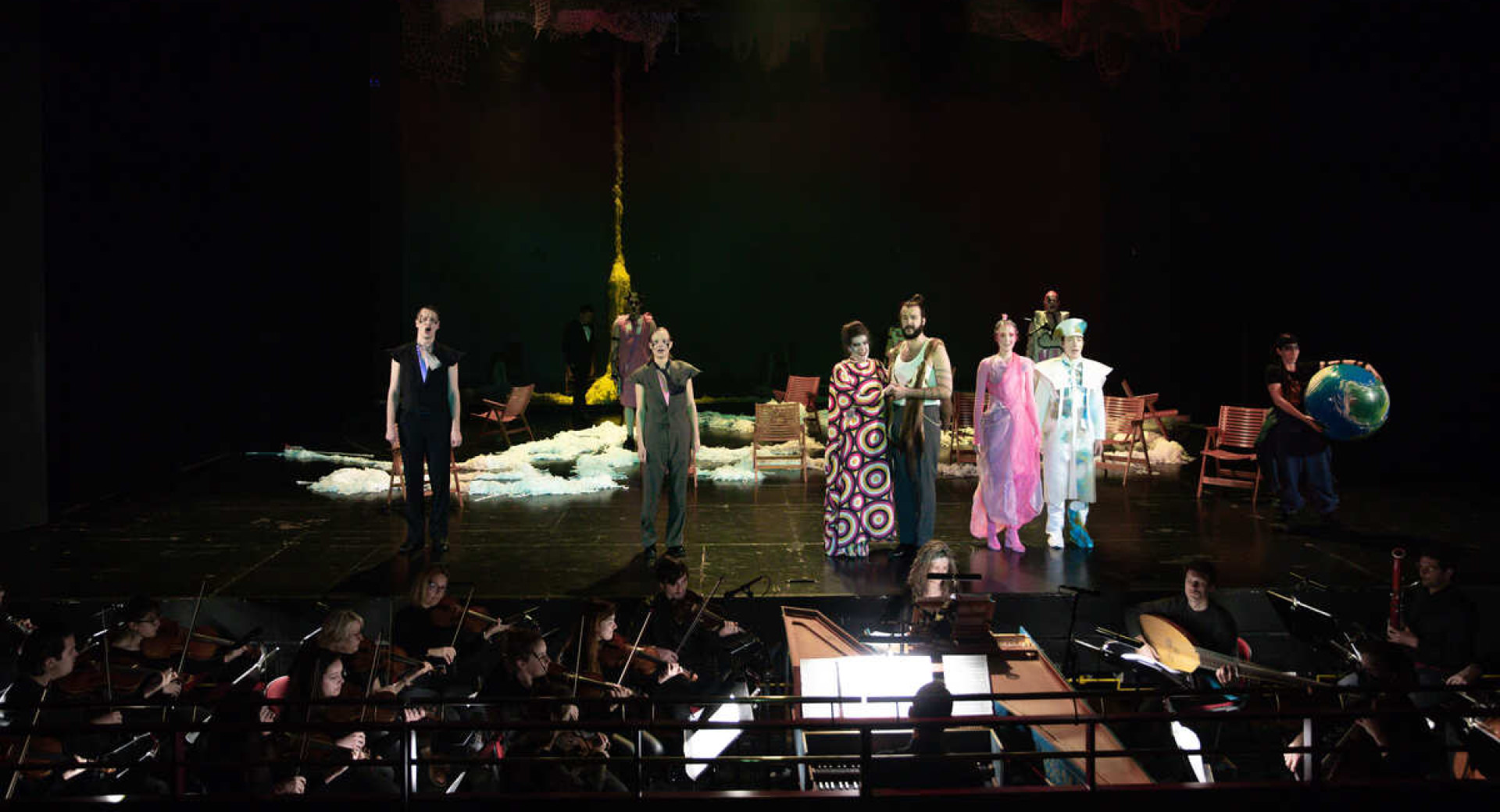 The actors stand on stage in costume, with the orchestra playing beneath the stage.
