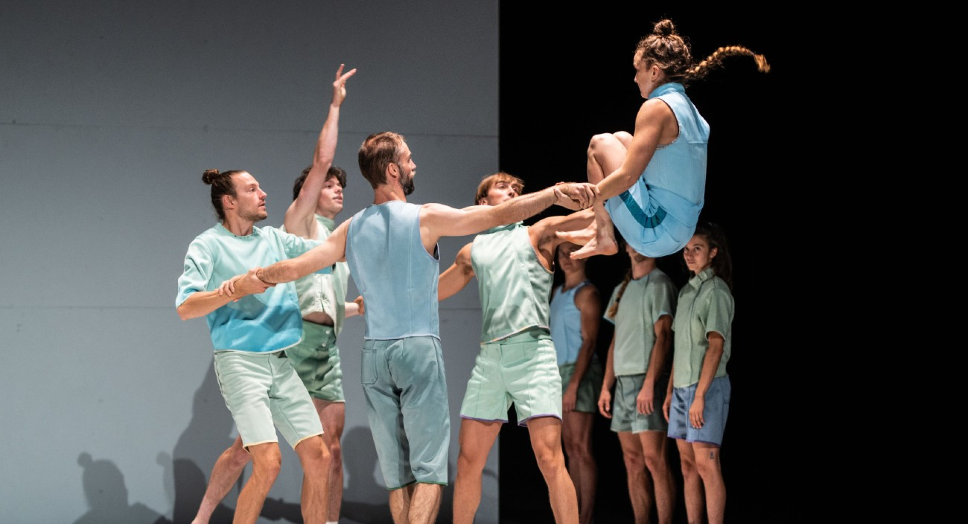 Osem plesalcev v pastelno zelenih in modrih oblacilih nastopa na odru; ena zenska je v zraku s pokrcenimi nogami, dva moska pa jo drzita za roke.