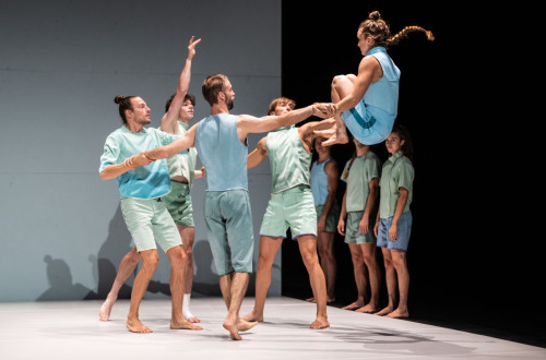 Osem plesalcev v pastelno zelenih in modrih oblacilih nastopa na odru; ena zenska je v zraku s pokrcenimi nogami, dva moska pa jo drzita za roke.