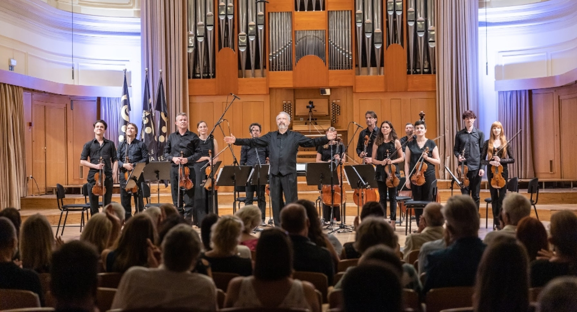 Dirigent in orkester stojijo na odru, v ospredju publika, v ozadju pa velike orgle.