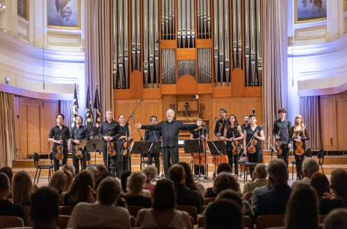 Dirigent in orkester stojijo na odru, v ospredju publika, v ozadju pa velike orgle.