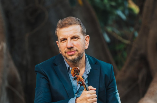 A man in an elegant teal suit is sitting on a big tree branch, holding a violin in his hands.