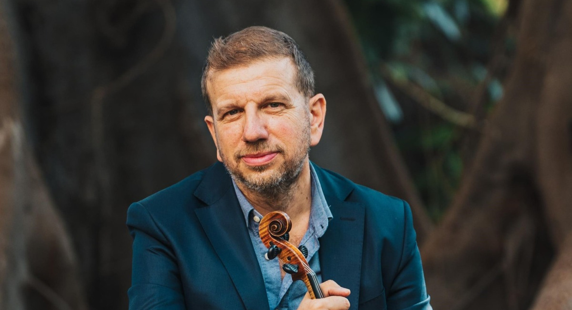 A man in an elegant teal suit is sitting on a big tree branch, holding a violin in his hands.
