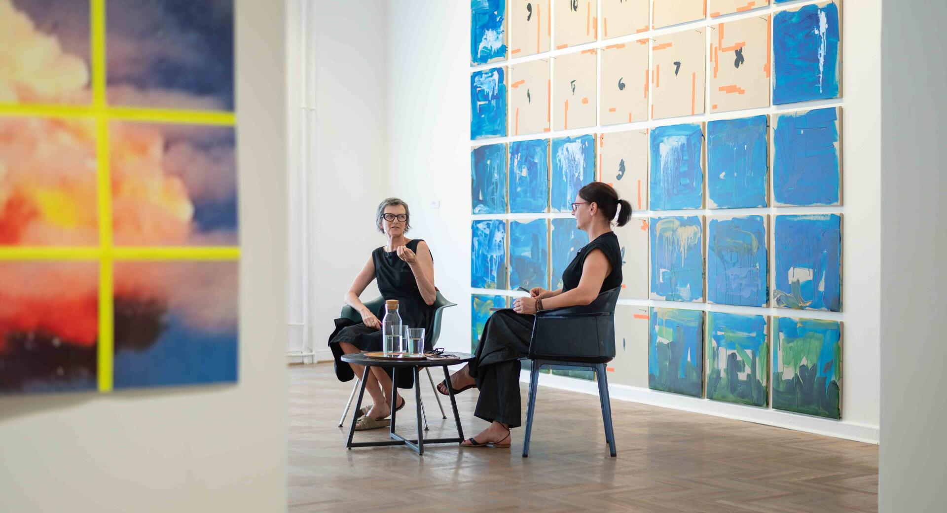 Dve ženski sedita in se pogovarjata v sodobni umetniški galeriji, v ozadju pa so razstavljena abstraktna umetniška dela v modrih, smetanastih in oranžnih odtenkih.