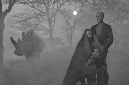 A black-and-white photograph of a devastated landscape with bare trees and a misty atmosphere; in the foreground, a dark-skinned couple with a baby, and in the background, two rhinoceroses.