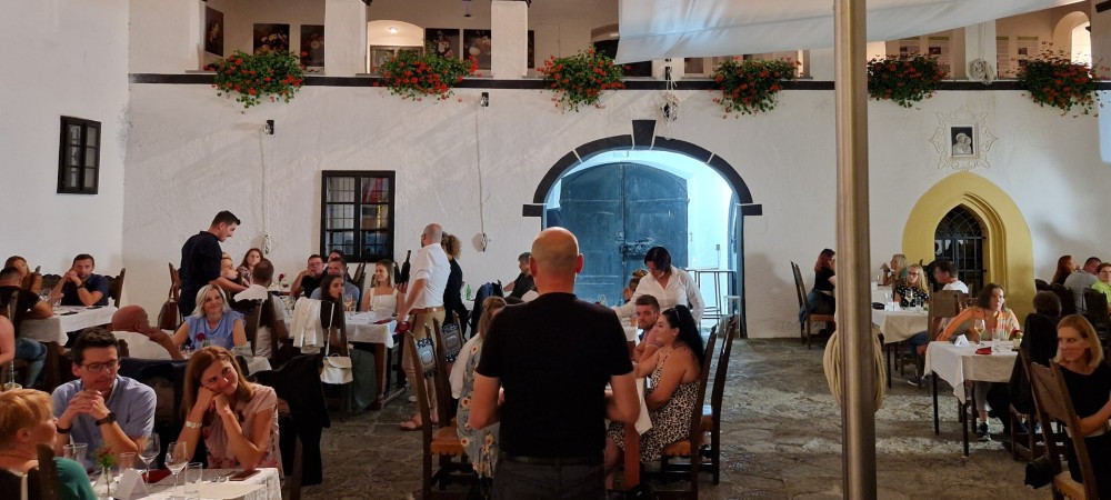 In the castle’s inner courtyard, people sit at tables, enjoying food and drink. Above them rises a balcony adorned with decorative flowers.