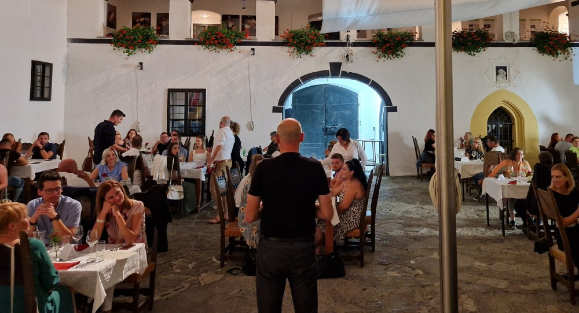 V grajskem notranjem atriju ljudje sedijo za mizami in uzivajo v hrani ter pijaci. Nad njimi se dviga balkon z okrasnimi cvetlicami.