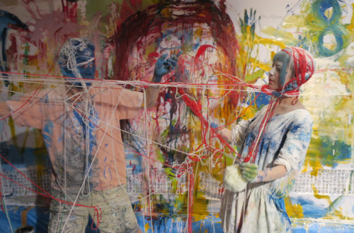 Dva cloveka v oblekah, poslikanih z barvami, odeta v pletenino iz raznobarvnih prejej, v ozadju pa platno, poslikano z razlicnimi barvami.
