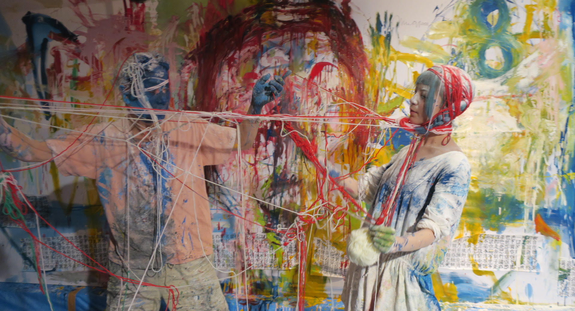 Two people wearing painted outfits, dressed in knitwear made of multicolored yarns, with a canvas painted in various colors in the background.