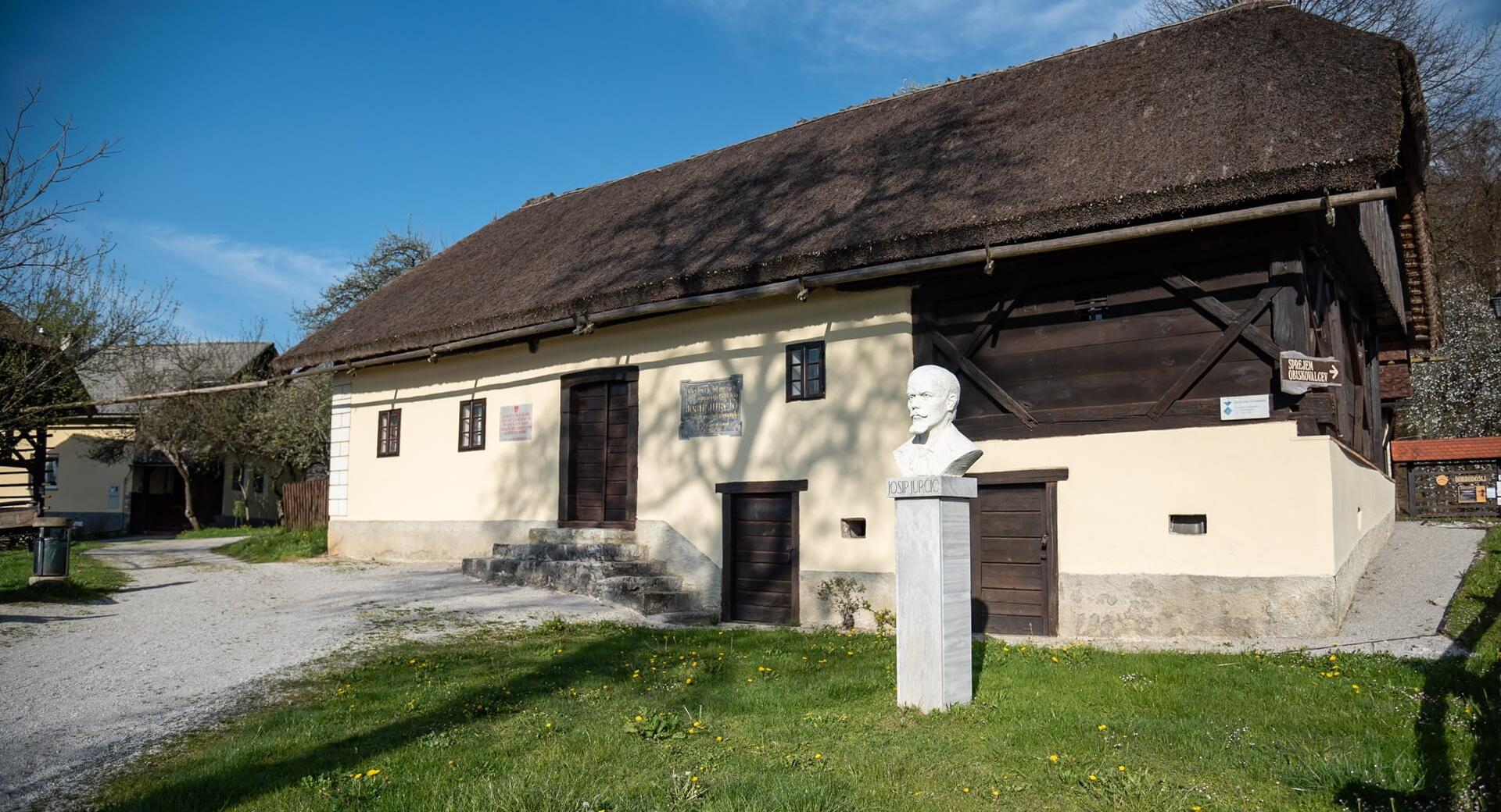 Tradicionalna hiša s slamnato streho, svetlimi ometanimi stenami in lesenimi vrati, pred katero na travniku stoji doprsni kip Josipa Jurčiča.