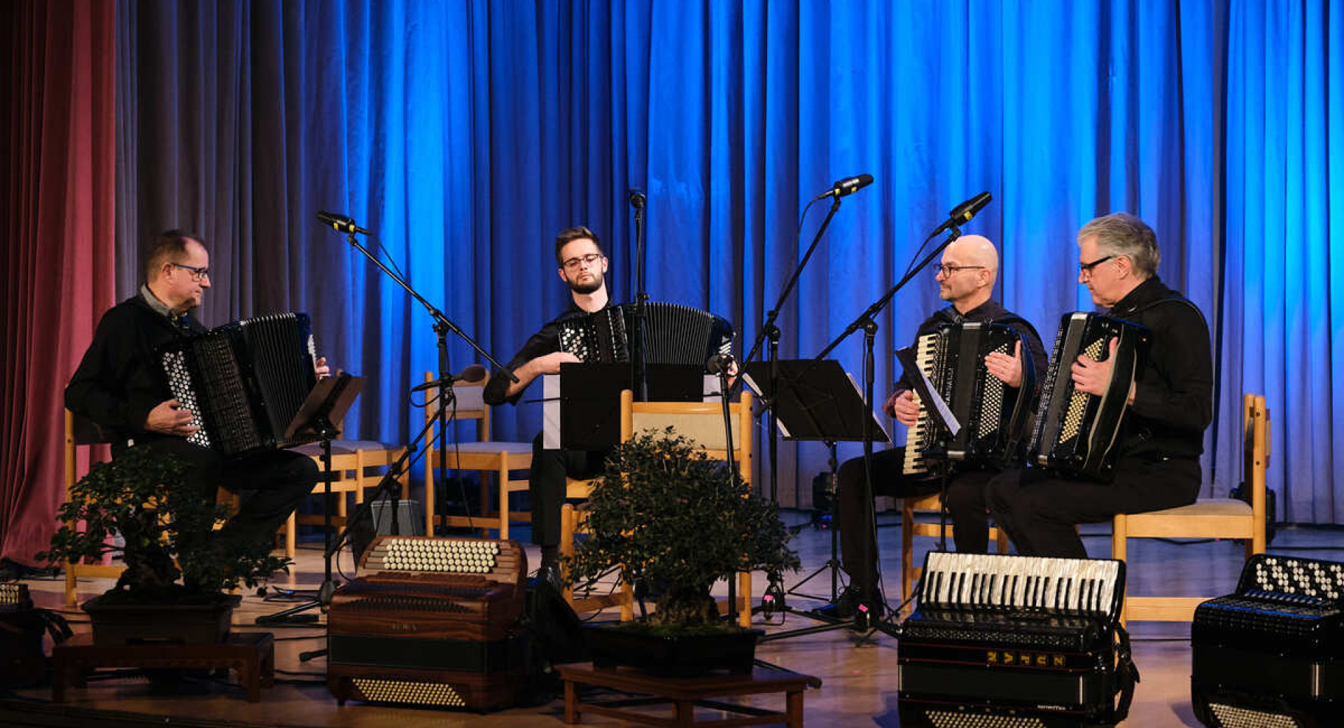 Four accordionists are seated on chairs on stage, playing their instruments, with a blue curtain backdrop behind them.