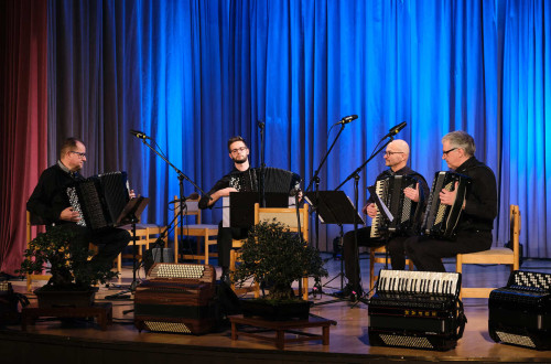 Four accordionists are seated on chairs on stage, playing their instruments, with a blue curtain backdrop behind them.