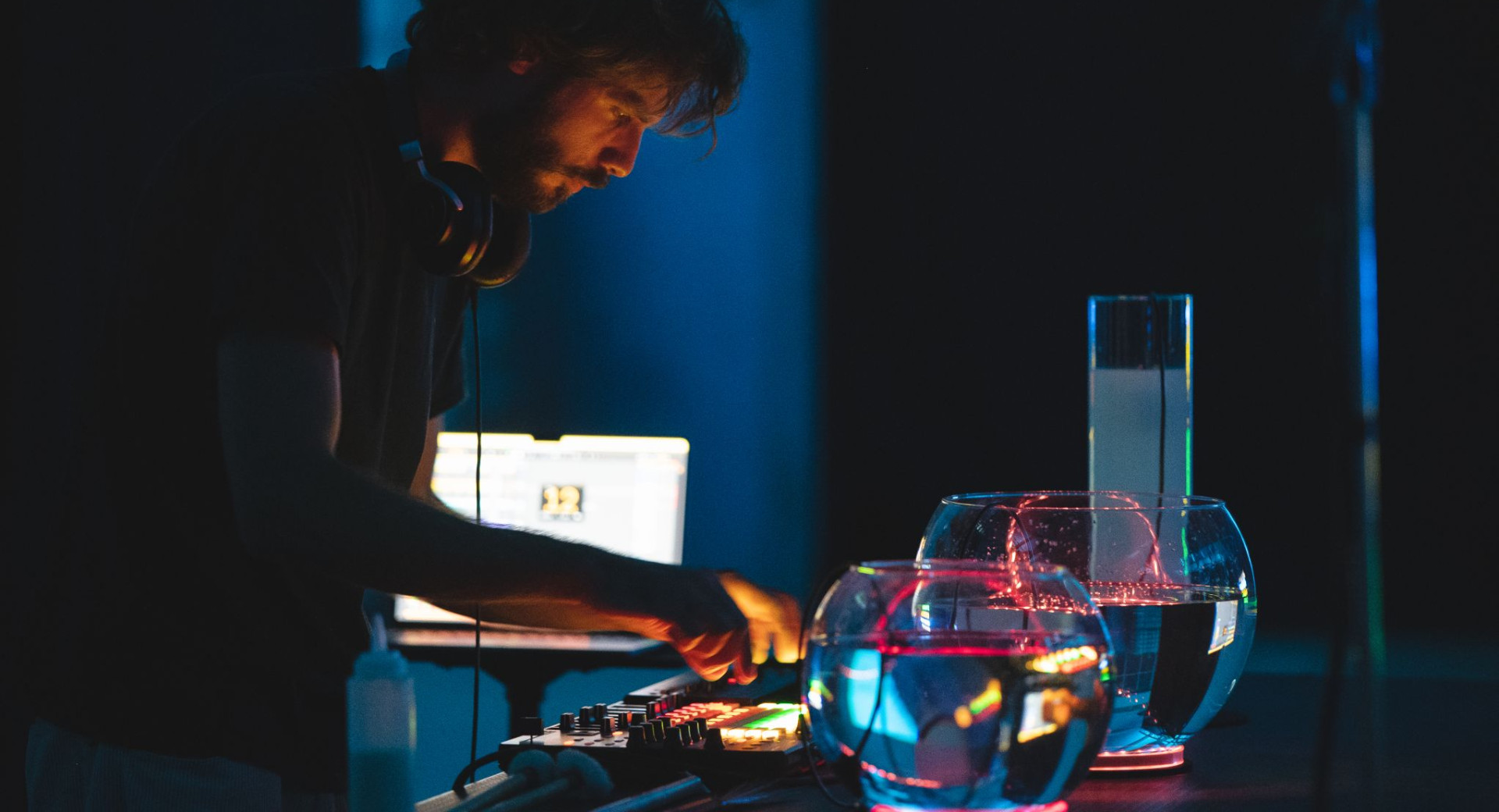 A man with headphones around his neck is mixing music on a mixing table, next to which are containers of water illuminated by coloured lights.
