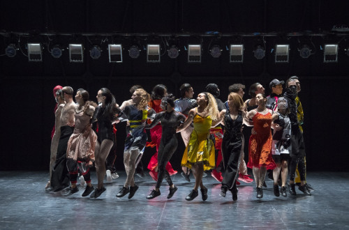 Skupina plesalcev v pisanih oblacilih plese v krogu na odru, kjer so obrnjeni navzven.