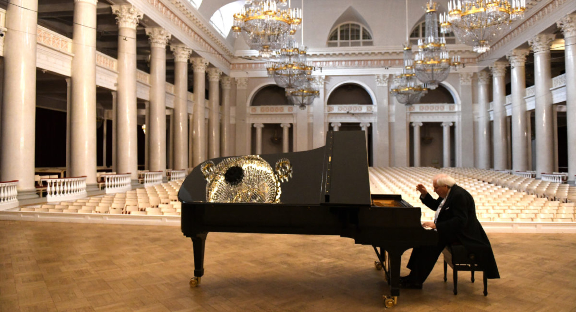 Pianist sedi in igra na crnem klavirju v razkosni dvorani z belimi stebri in zlatimi lestenci.