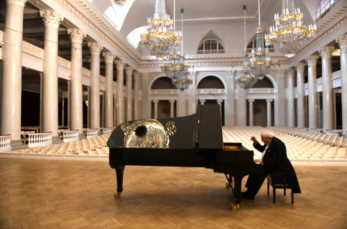 Pianist sedi in igra na crnem klavirju v razkosni dvorani z belimi stebri in zlatimi lestenci.