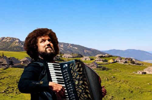 A man is playing the accordion in front of a green landscape with wooden houses and mountains in the background.