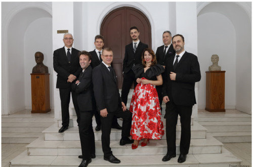 Skupina sedmih moskih in ene zenske v elegantnih oblacilih stoji na belih stopnicah pred velikimi lesenimi vrati.