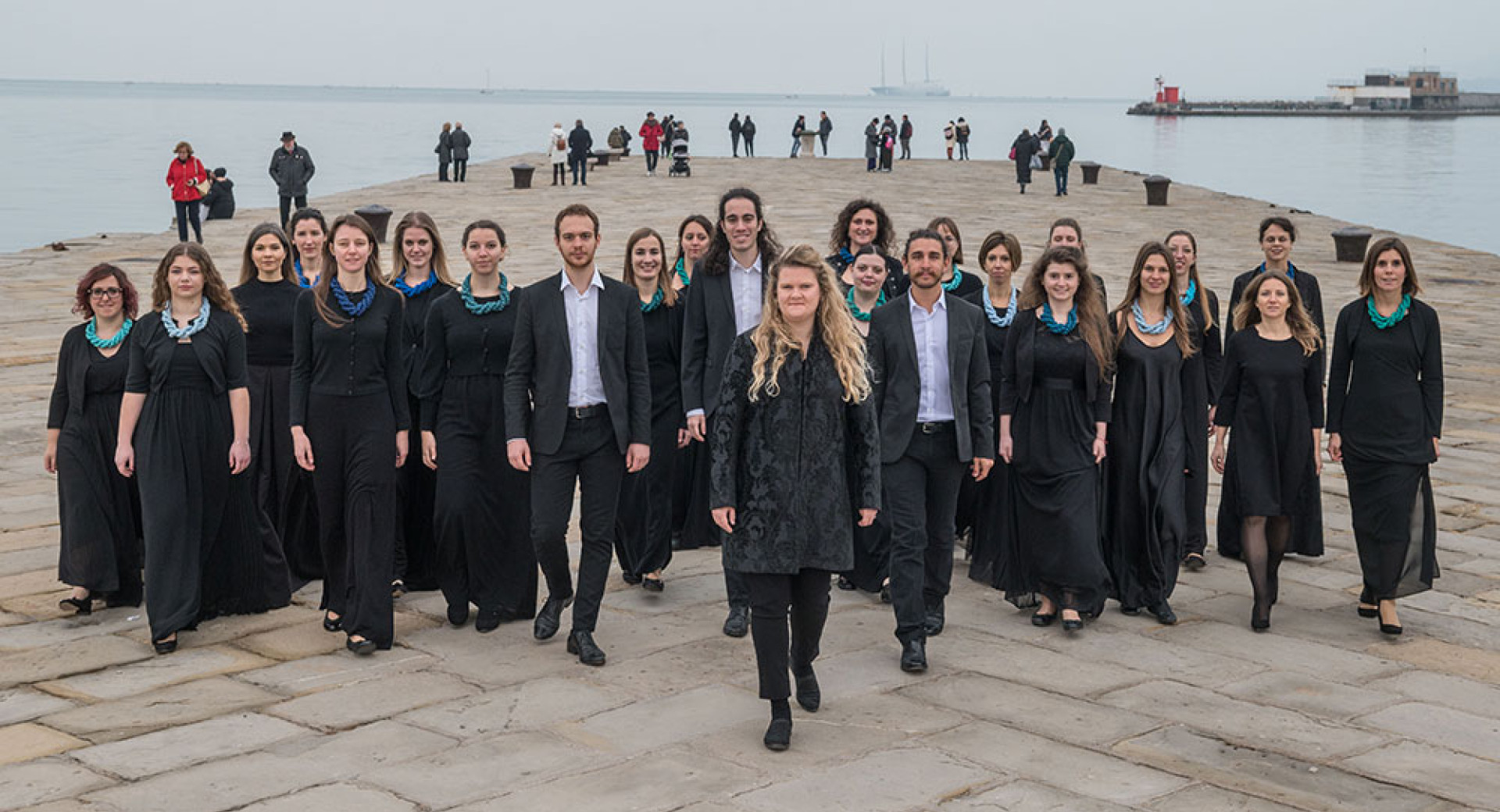 A group of people in black clothes standing on a large pier with the sea in the background.