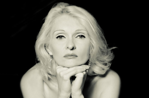 Crno-bela fotografija zenske s svetlimi lasmi, ki sedi z glavo naslonjeno na roke.