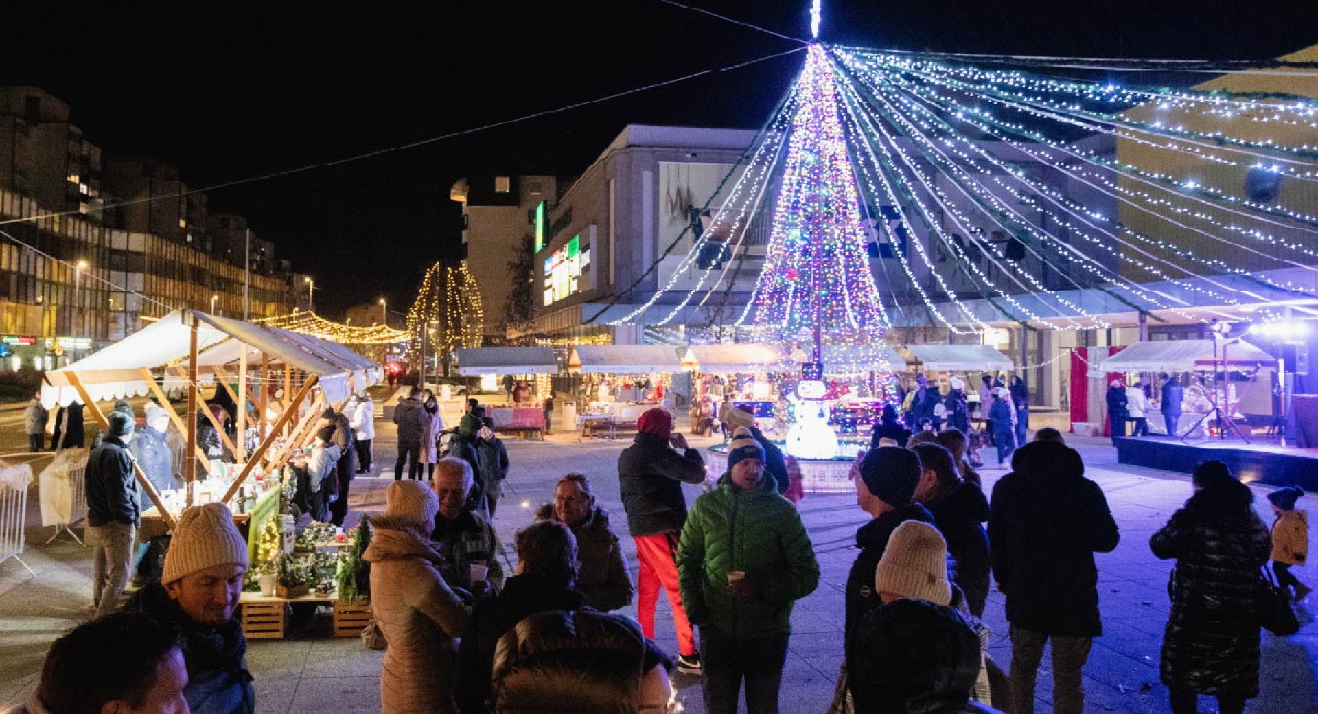 Toplo obleceni ljudje so zbrani na prostem v mraku, okoli njih pa stojnice in drsalisce, praznicno okraseni z luckami.