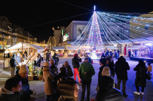 Toplo obleceni ljudje so zbrani na prostem v mraku, okoli njih pa stojnice in drsalisce, praznicno okraseni z luckami.