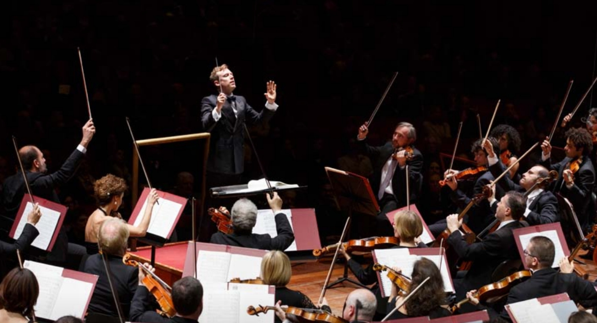 The conductor conducts the orchestra, where violinists with their bows raised in the air are visible.