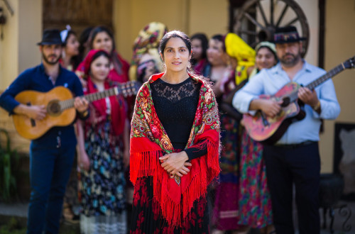 Zenska v crnem z rdeco ruto okoli ramen in clani skupine v ozadju.