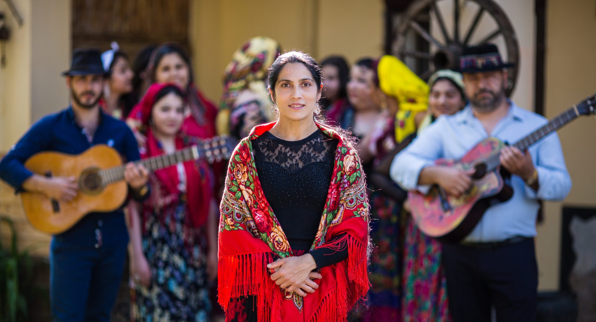 Zenska v crnem z rdeco ruto okoli ramen in clani skupine v ozadju.