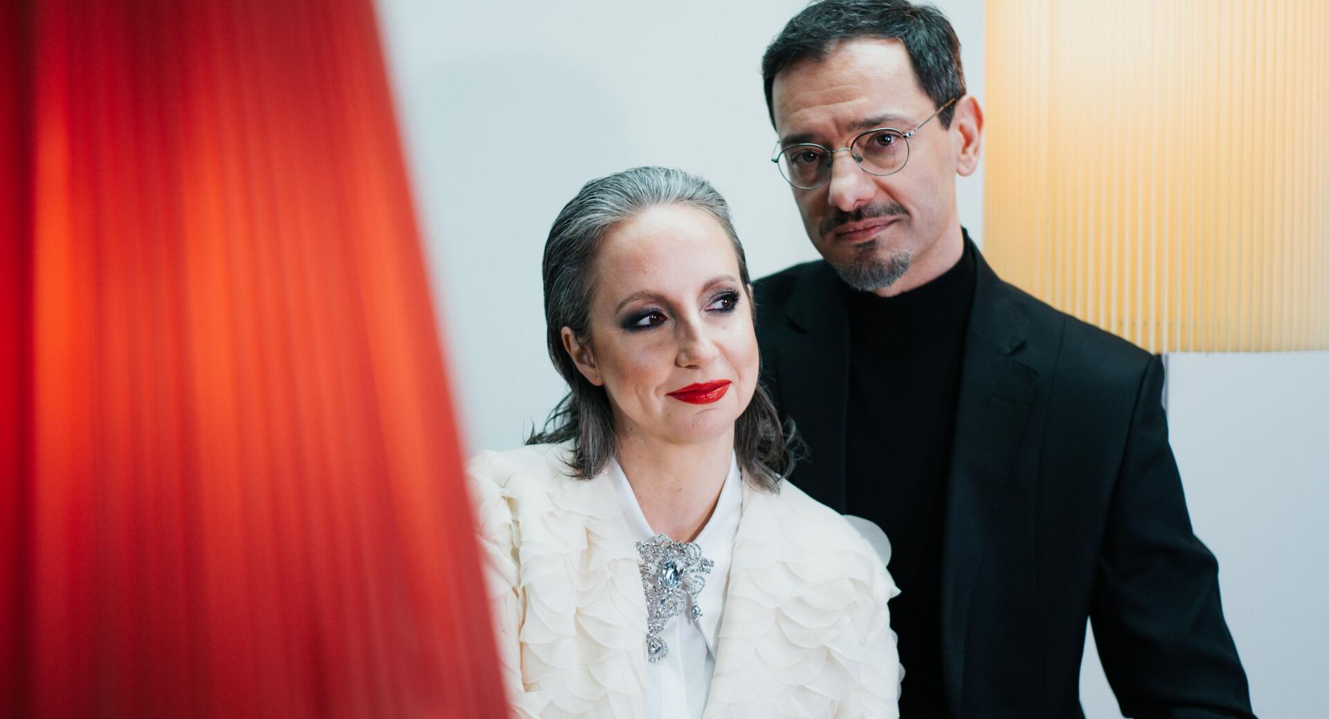 Portret prikazuje žensko s sivimi lasmi, izrazito ličili in v elegantnem belem suknjiču z broško ter moškega z očali in v črnem sakoju, ki stoji za njo, medtem ko je v levem ospredju zamegljen rdeč element.