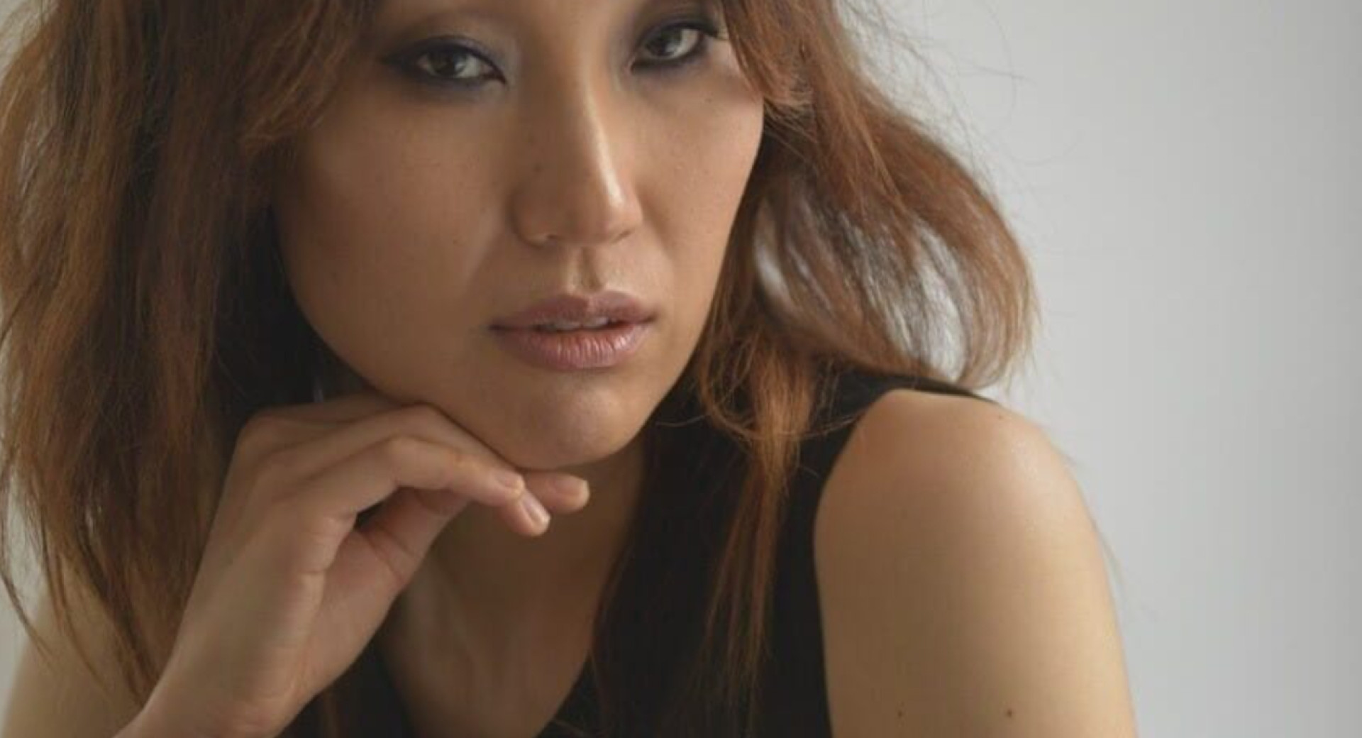 A portrait of a woman with brown hair in a black top, sitting with her head supported by her right hand under her chin.