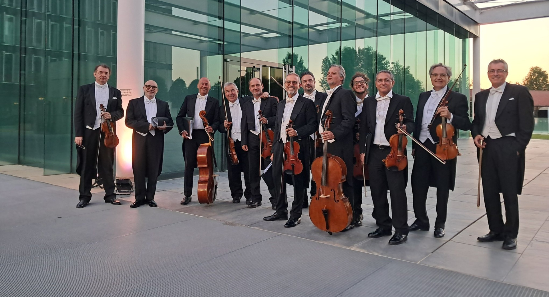 Men with instruments are standing outside on a stone platform in front of a glass building.