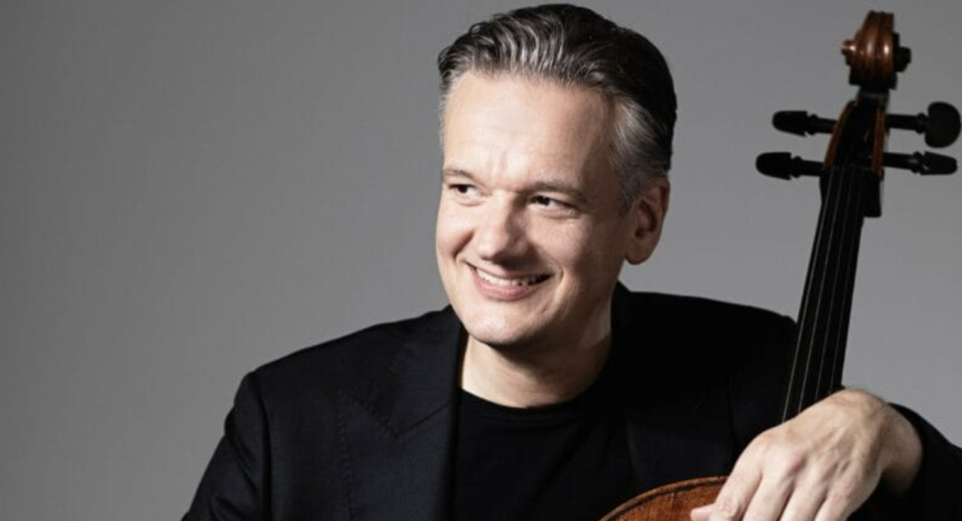Moski v crnem suknjicu sedi, gleda v desno in z levo roko objema violoncelo.
