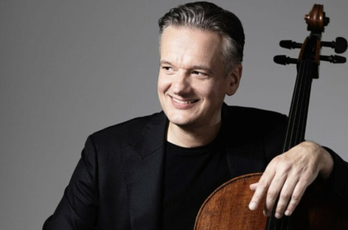 Moski v crnem suknjicu sedi, gleda v desno in z levo roko objema violoncelo.