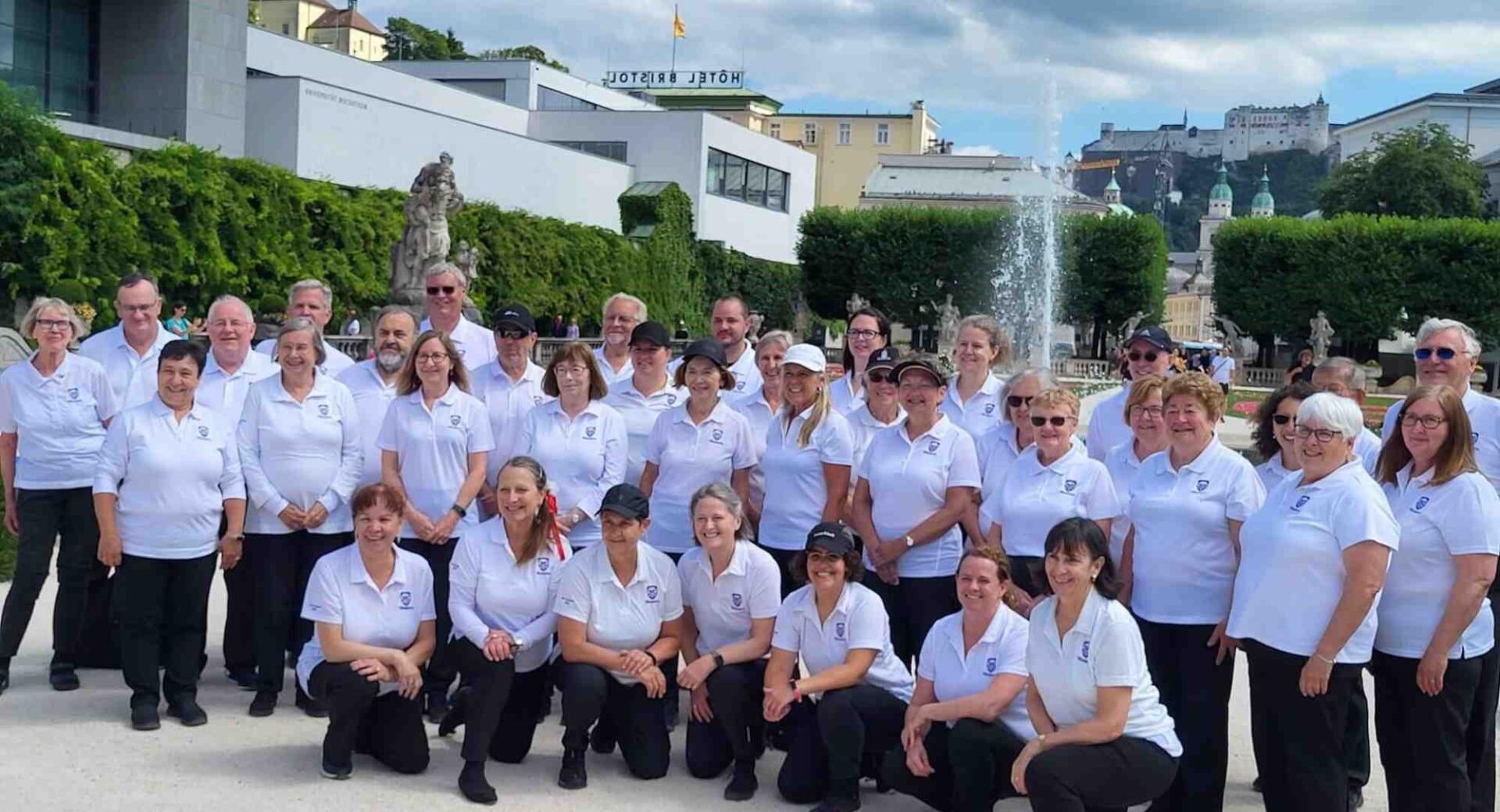 Skupinska fotografija večje skupine ljudi v ujemajočih se belih polo majicah in črnih hlačah, ki pozirajo na prostem v parku s fontano in zgodovinsko mestno kuliso v ozadju.