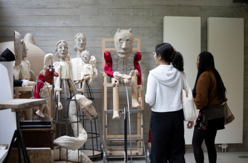Three women are viewing an exhibition of various sculptures made from different materials.