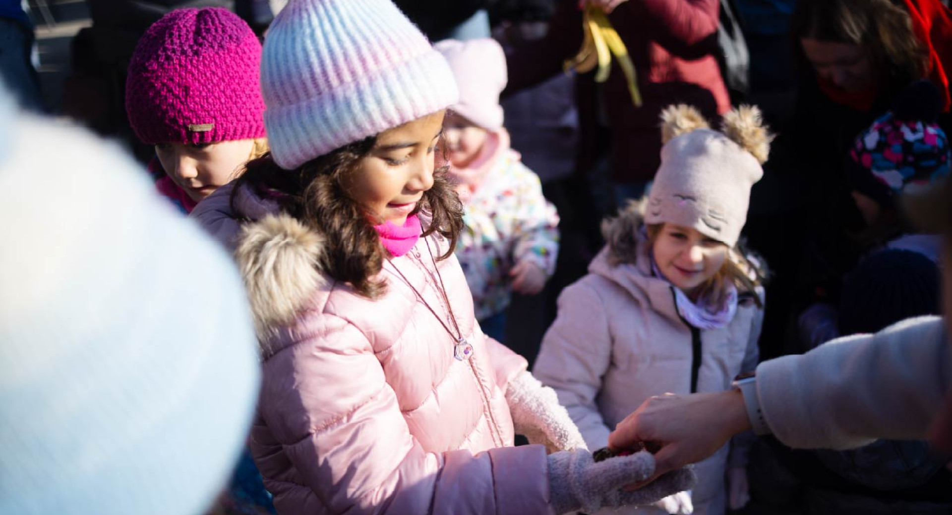 Mlada punca v roza bundi, kapi in rokavicah prejema darilo, v ozadju pa je se vec otrok.