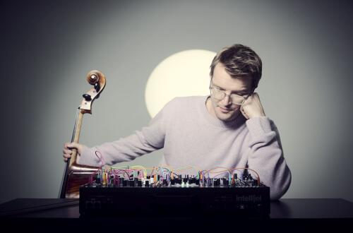 Moški sedi z violončelom v roki in elektronsko avdio opremo na črni mizi, naslonjen z glavo na roko v mehko osvetljeni sobi.