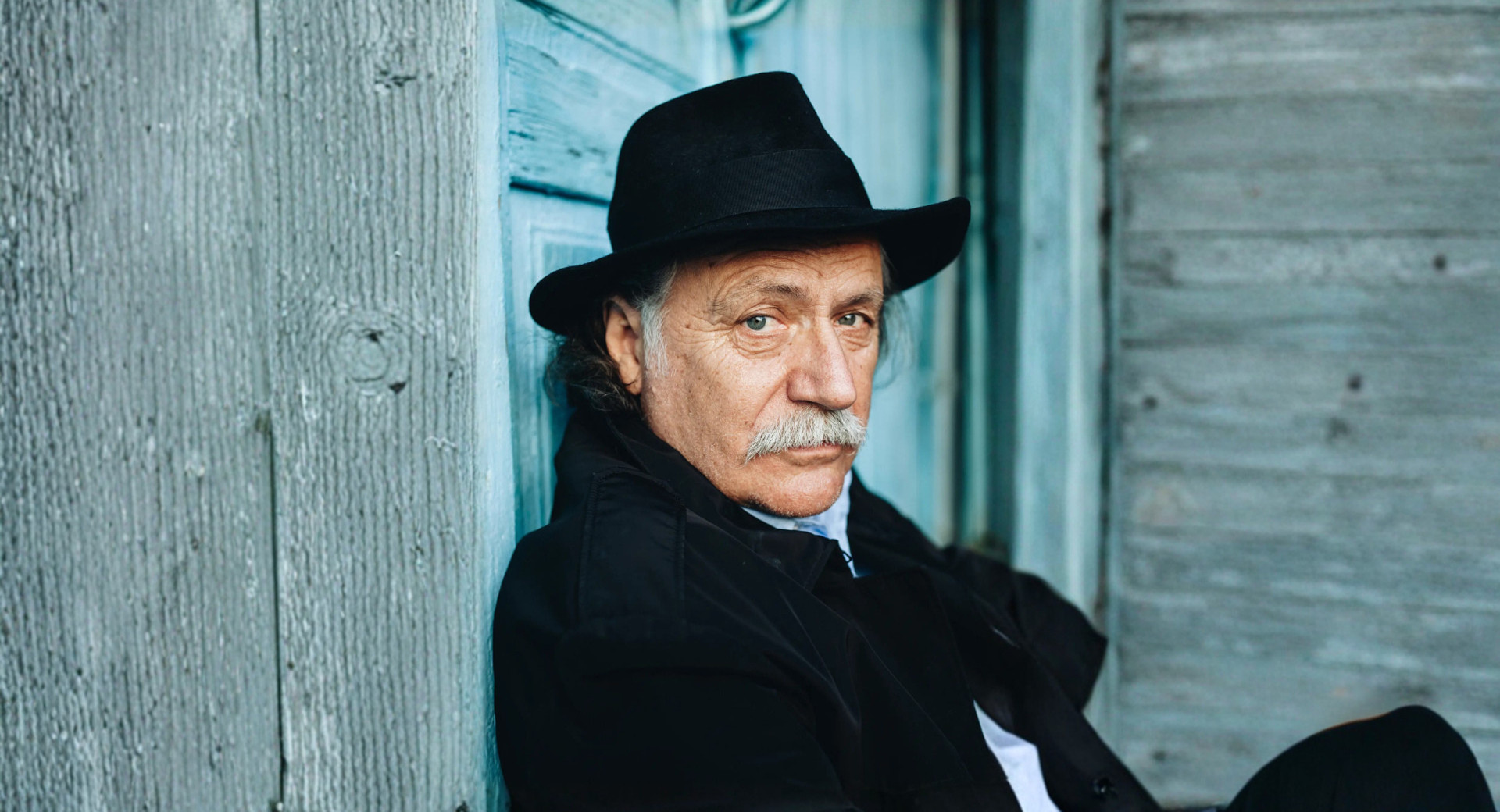 Starejsi moski s klobukom v crnih oblacilih zre v objektiv.