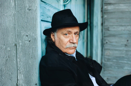 Starejsi moski s klobukom v crnih oblacilih zre v objektiv.