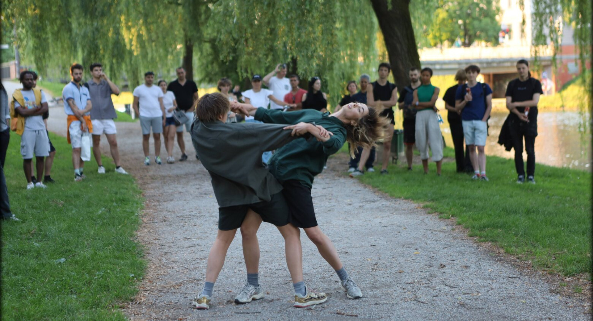 Two dancers in sportswear are dancing on a public surface, on a sandy path surrounded by grass, while spectators watch them in the background.